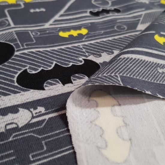 Tela Punto Algodón Batman Logos - Tela de punto algodón licencia con dibujos de logos de Batman en varios colores y tamaños sobre un fondo donde predominan los colores grises. La tela mide 150cm de ancho y su composición 92% aglodón – 8% elastano.